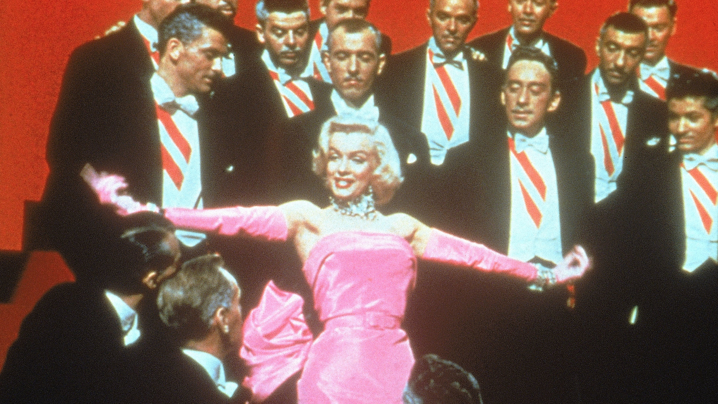 "Blondinen bevorzugt": Eine attraktive blonde Showtänzerin steht auf einer Bühne umringt von sie anhimmelnden Männern in schwarzem Frack, rot-weiß-gestreifter Schärpe und mit grauen Schläfen. Die Frau trägt ein leuchtend pinkes, schulterfreies, langes Abendkleid aus Taft mit passenden achselhohen Handschuhen und aufwändigem Diamantschmuck.