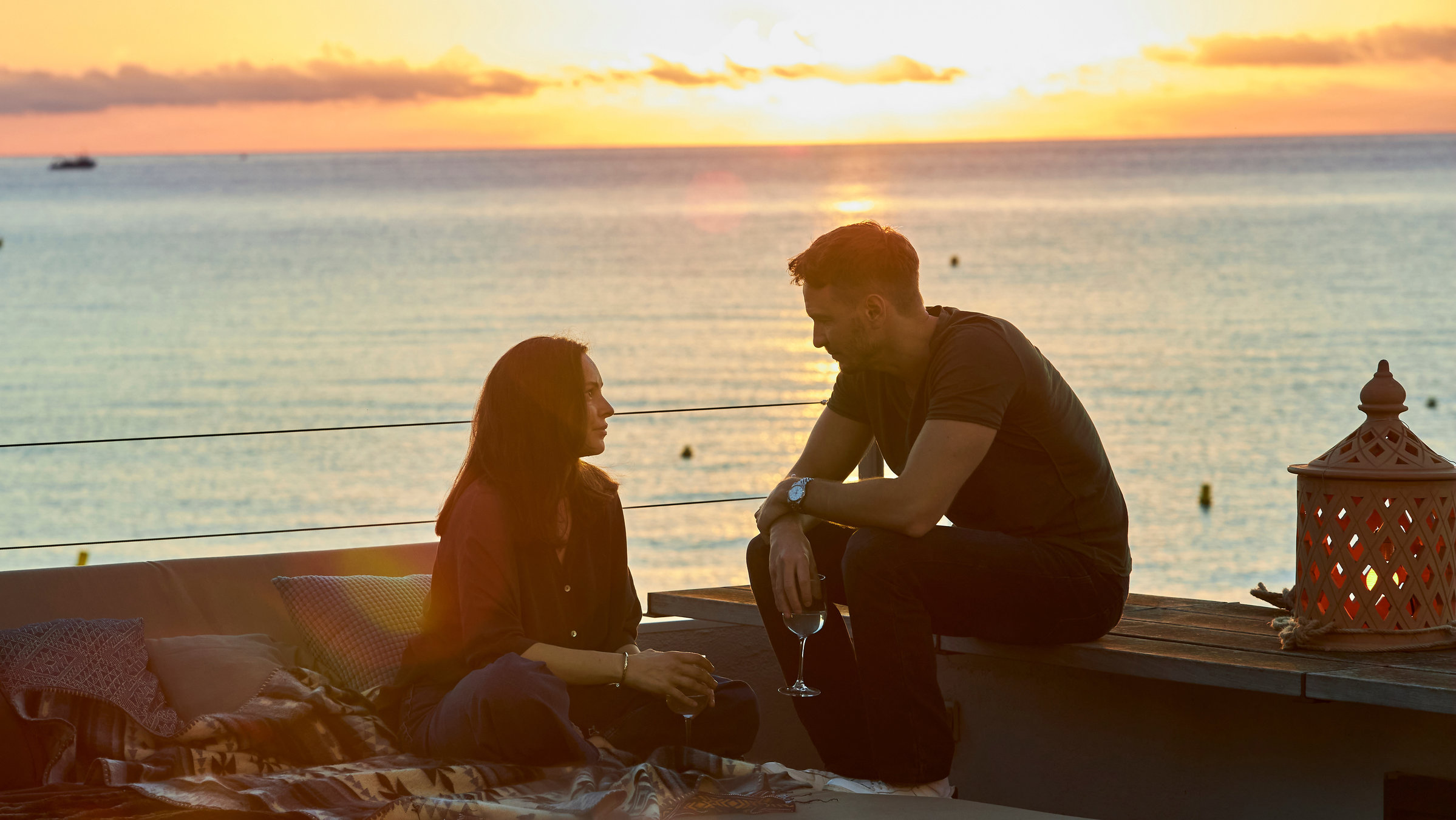 "Hotel Barcelona - Teil 2": Laura Santos (Mina Tander) und Mateo Bonachera (Vladimir Burlakov) sitzen mit einem Glas Wein in der Hand auf der Dachterrasse seiner Wohnung und schauen sich an. Hinter ihnen liegt das Meer bei Sonnenuntergang.