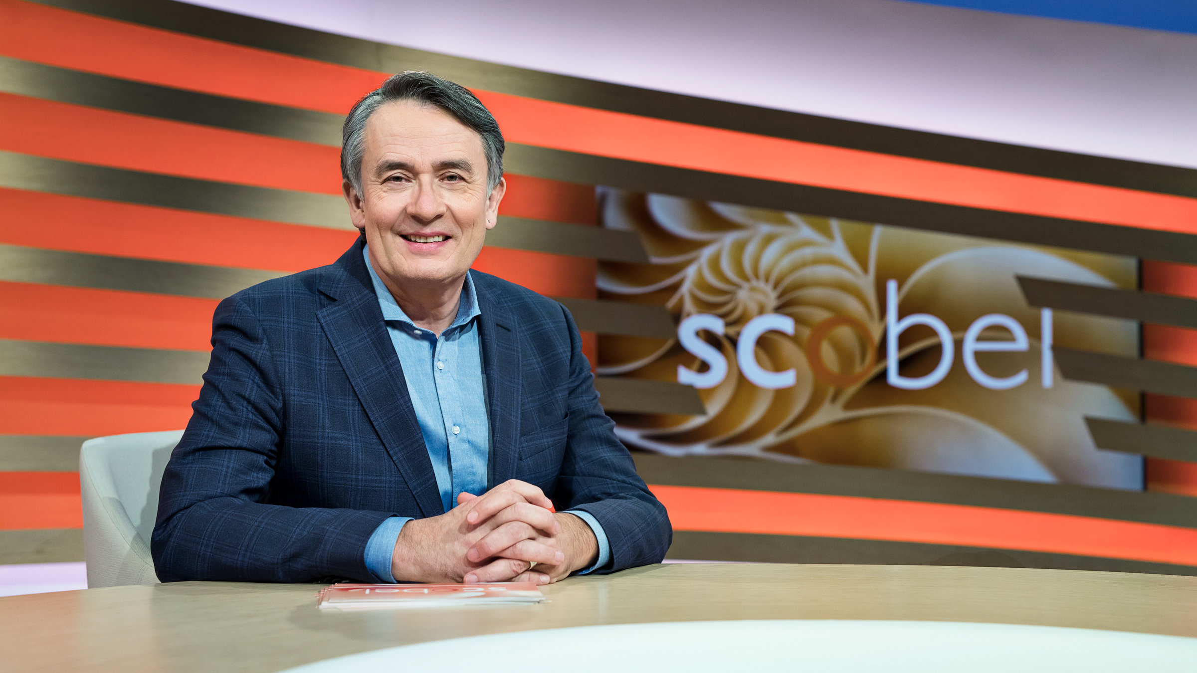 "scobel": Gert Scobel sitzend am Tisch in Studio-Deko.