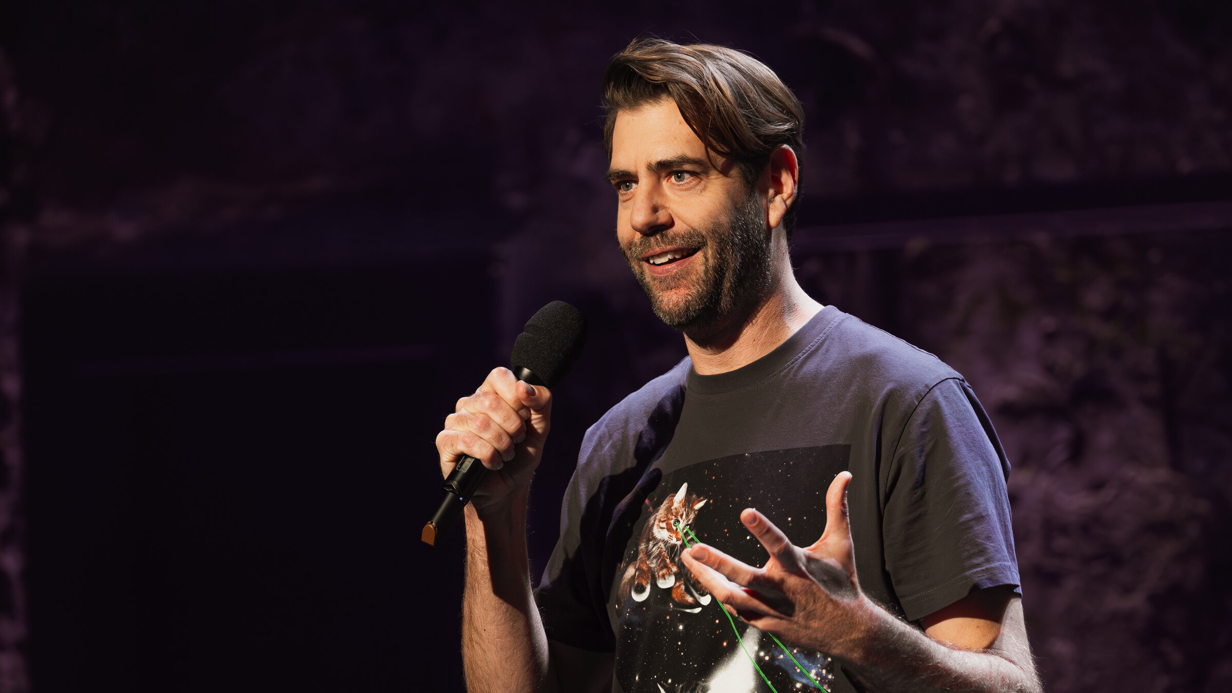 "Till Reiners´ Happy Hour - Takeover": Der Kabarettist Claus von Wagner, der in dieser Spezialfolge die Moderation der Sendung übernimmt, steht mit einem Mikrofon in der Hand auf der Bühne und lächelt Richtung Publikum.