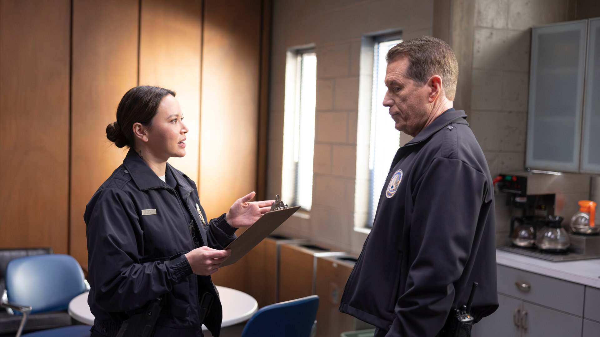 "The Rookie - Babysitting": Lucy (Melissa O'Neil) steht Smitty (Brent Huff) in einem Pausenraum gegenüber. Ihr Blick ist fragend und zugleich streng, während sie ein Klemmbrett in der Hand hält.