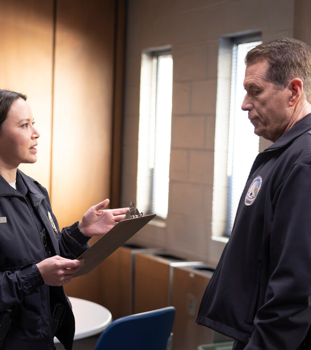 "The Rookie - Babysitting": Lucy (Melissa O'Neil) steht Smitty (Brent Huff) in einem Pausenraum gegenüber. Ihr Blick ist fragend und zugleich streng, während sie ein Klemmbrett in der Hand hält.