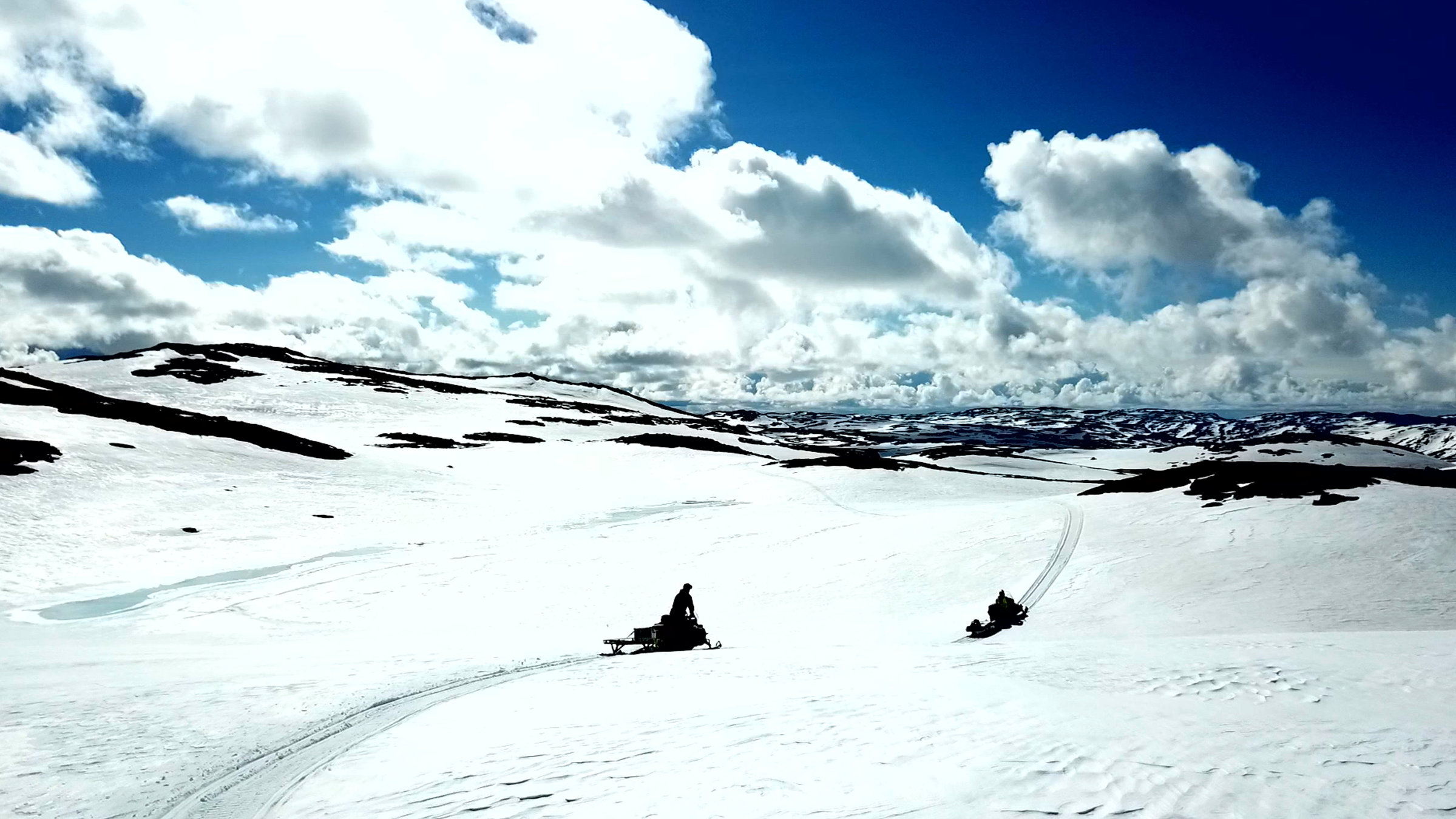 "Skandinaviens versteckte Paradiese": Totale: Zwei Schneemobile fahren von rechts nach links über weite Schneeflächen, Himmel dunkelblau mit weißen Wolken.