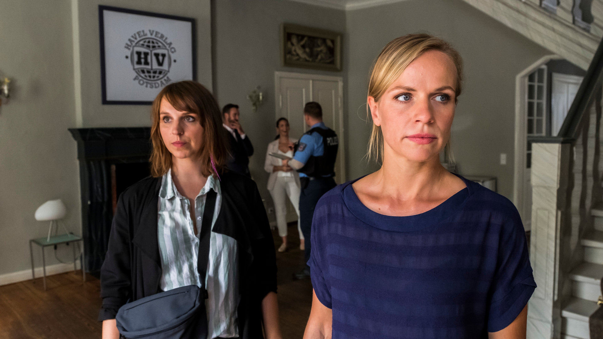 "SOKO Potsdam - Küsse in St. Tropez": Luna Kunath (Caroline Erikson) steht hinter Sophie Pohlmann (Katrin Jaehne) im Eingangsbereich einer Villa. Die Kommissarinnen blicken beide mit nachdenklichem Gesichtsausdruck zur Seite. Im Hintergrund sieht man einen Mann und eine Frau mit einem Polizisten sprechen. Außerdem hängt an der Wand ein eingerahmtes Bild auf dem das Logo "Havel Verlag Potsdam" zu sehen ist.