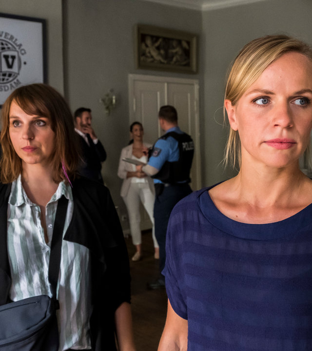 "SOKO Potsdam - Küsse in St. Tropez": Luna Kunath (Caroline Erikson) steht hinter Sophie Pohlmann (Katrin Jaehne) im Eingangsbereich einer Villa. Die Kommissarinnen blicken beide mit nachdenklichem Gesichtsausdruck zur Seite. Im Hintergrund sieht man einen Mann und eine Frau mit einem Polizisten sprechen. Außerdem hängt an der Wand ein eingerahmtes Bild auf dem das Logo "Havel Verlag Potsdam" zu sehen ist.