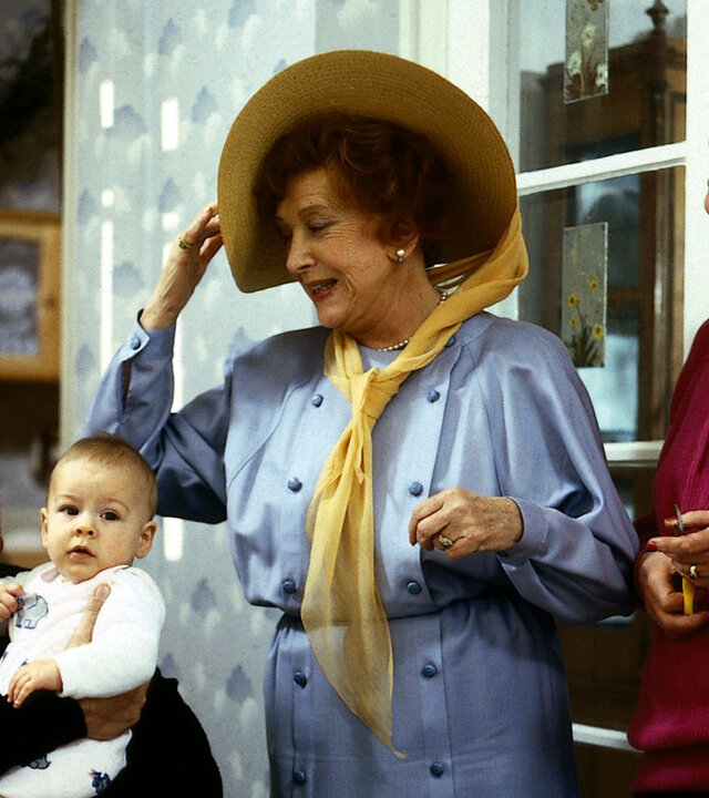"Die Wicherts von nebenan: Ein Auto für Sizilien": Eberhard Wichert (Stefan Orlac) sitzt mit dem Baby auf dem Arm im Wohnzimmer. Oma Käthe (Edith Schollwer) und Hannelore (Maria Sebaldt) stehen neben ihm. Oma Käthe trägt einen Hut.