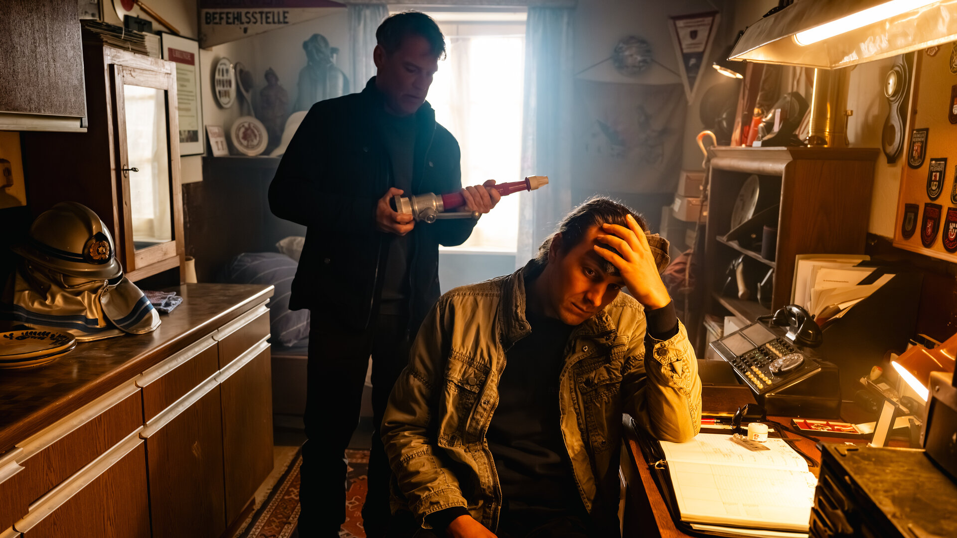 "SOKO Köln - Feuer und Flamme": Nils Steiner (Frederik Götz) sitzt an einem braunen Schreibtisch und hat den Kopf in seine linke Hand gestützt. Er sitzt in Richtung des Betrachters und blickt traurig nach unten. Hinter ihm steht Matti Wagner (Pierre Besson) und hält einen länglichen Gegenstand in den Händen, den er interessiert betrachtet. Eine Lampe über dem Schreibtisch spendet warmes Licht. Durch ein Fenster hinter Matti fällt Tageslicht in den Raum.