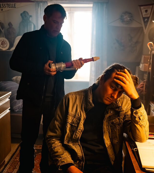 "SOKO Köln - Feuer und Flamme": Nils Steiner (Frederik Götz) sitzt an einem braunen Schreibtisch und hat den Kopf in seine linke Hand gestützt. Er sitzt in Richtung des Betrachters und blickt traurig nach unten. Hinter ihm steht Matti Wagner (Pierre Besson) und hält einen länglichen Gegenstand in den Händen, den er interessiert betrachtet. Eine Lampe über dem Schreibtisch spendet warmes Licht. Durch ein Fenster hinter Matti fällt Tageslicht in den Raum.