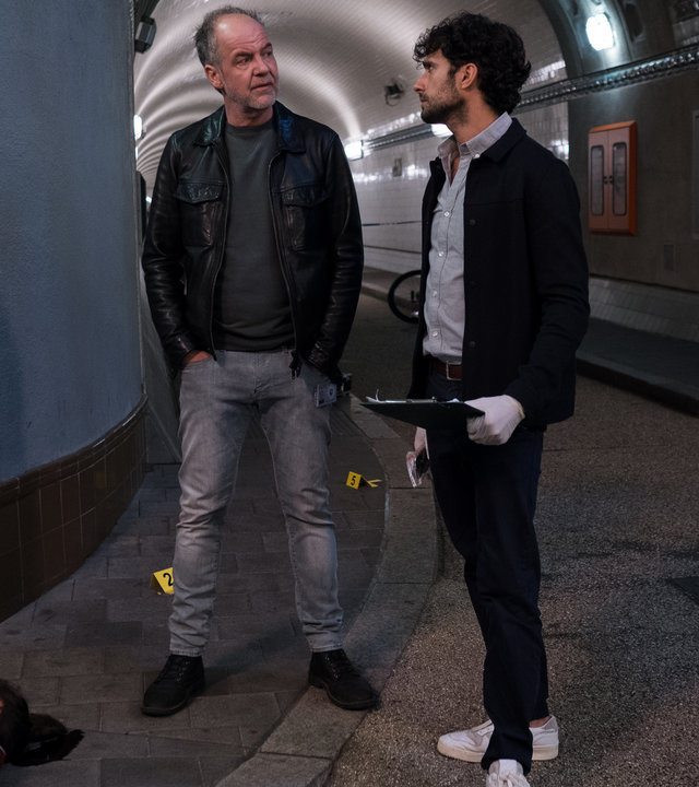 "SOKO Hamburg - Hafennixen": Oskar Schütz (Marek Erhardt) und Max Nordmann (Garry Fischmann) stehen in einer Unterführung vor der Leiche von Liv Thomsen (Maike Knirsch) und sehen sich nachdenklich an. Max hält ein schwarzes Klemmbrett in der Hand und trägt weiße Gummihandschuhe. Im Hintergrund sieht man den hell beleuchteten Tunnel der Unterführung. Der Bereich, in dem die Kommissare stehen, wird vom Licht ausgespart.