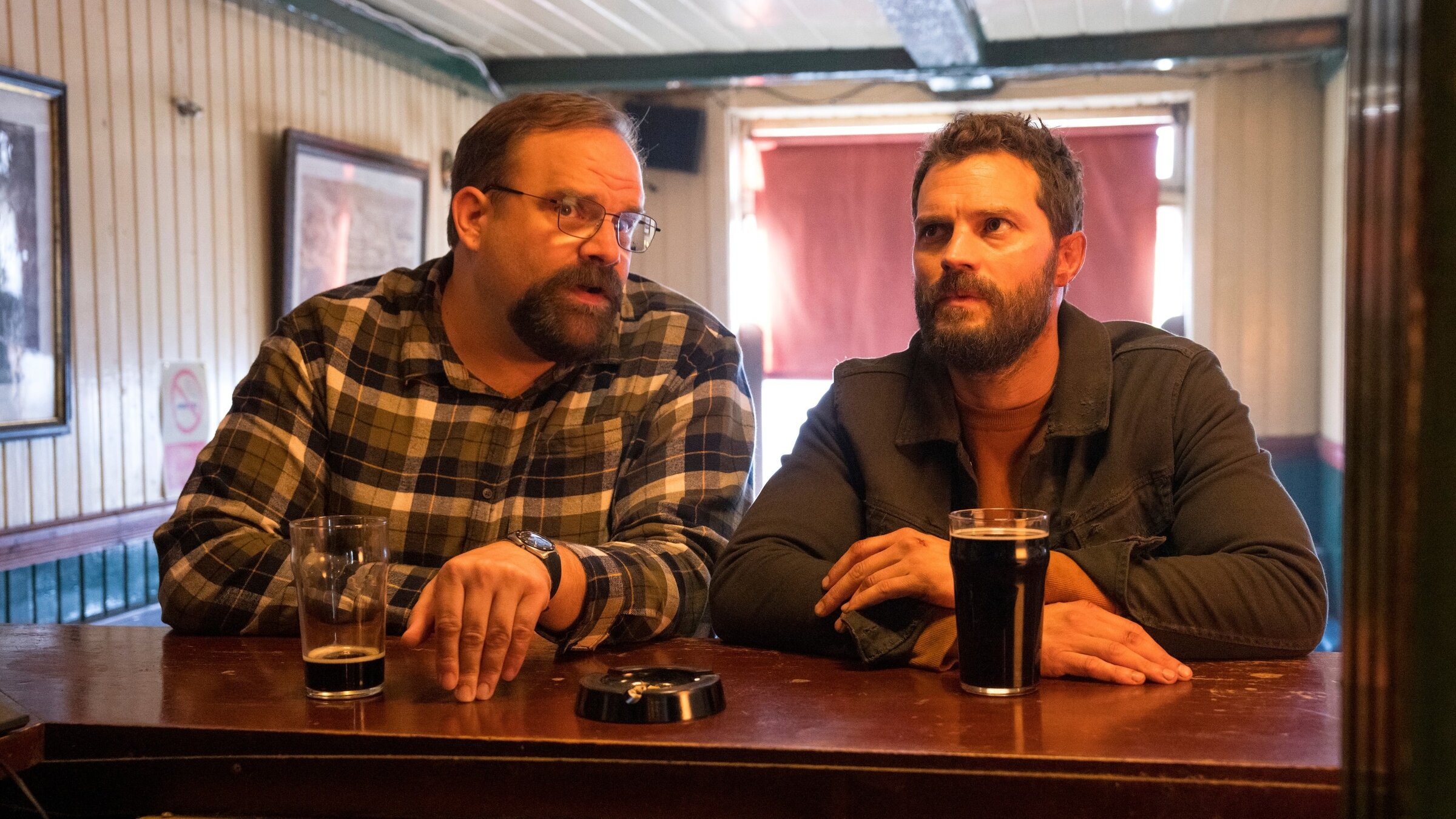 "The Tourist - Irisches Blut - Wurst und Musik": Ethan (Greg Larsen) und Elliot (Jamie Dornan) sitzen nebeneinander an einer Bar und unterhalten sich. Vor ihnen stehen dunkle Biere.