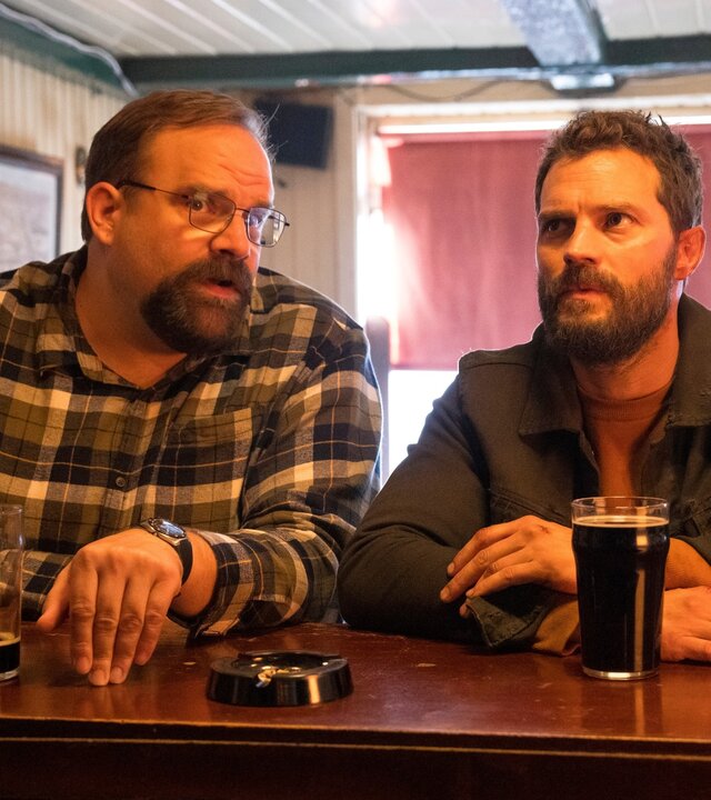 "The Tourist - Irisches Blut - Wurst und Musik": Ethan (Greg Larsen) und Elliot (Jamie Dornan) sitzen nebeneinander an einer Bar und unterhalten sich. Vor ihnen stehen dunkle Biere.