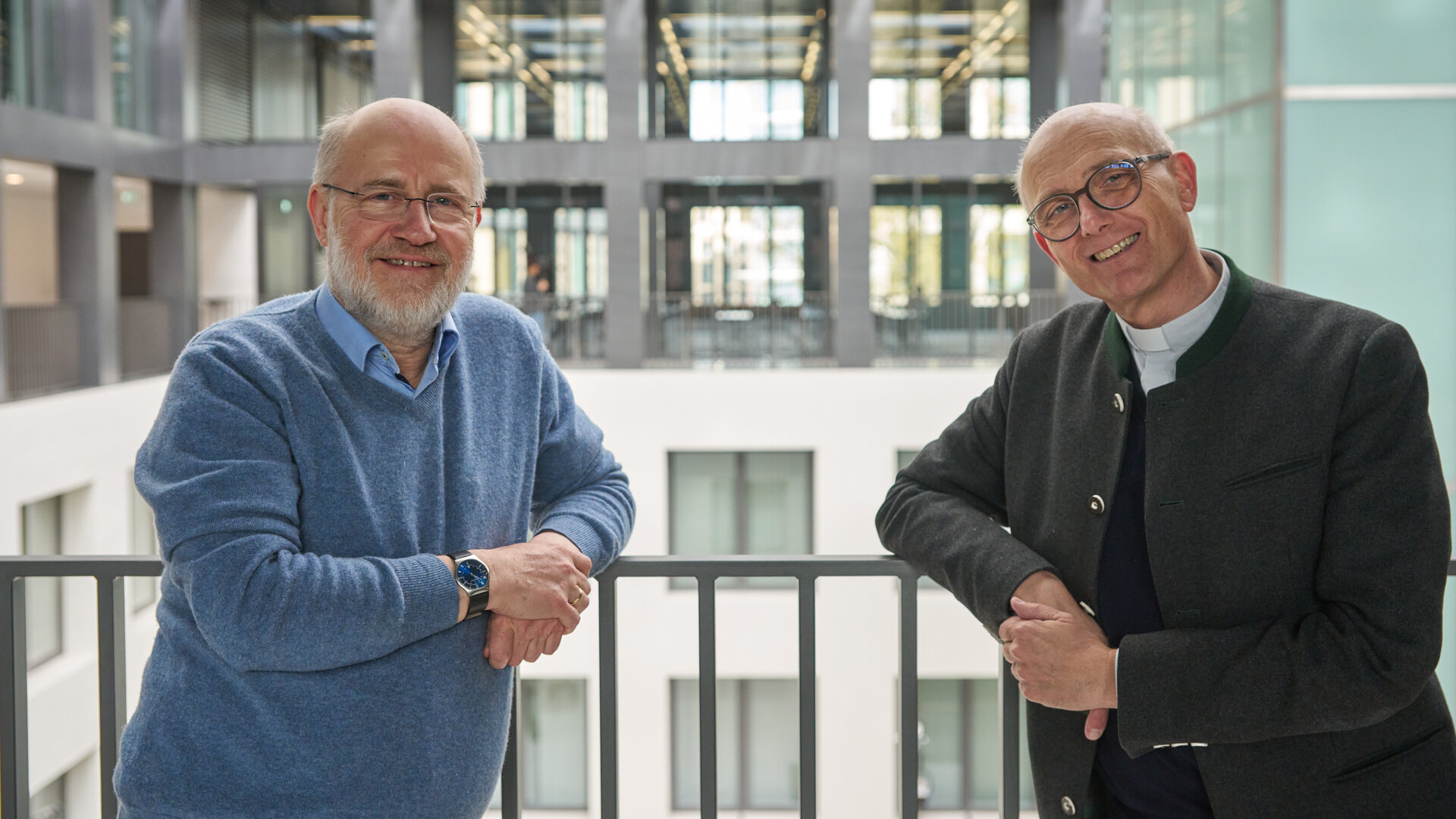 "Lesch sieht Schwartz: Solidarisch sein - was bringt's?": Harald Lesch und Thomas Schwartz stehen hoch oben in einem modernen Hochhaus vor einem Innenhof und lächeln in die Kamera.