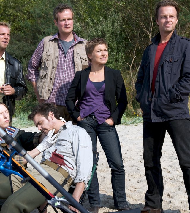 "SOKO Wismar - Haie und andere Hechte": In einem Campingstuhl am Strand sitzt zusammengesunken der tote Mikka Salminen (Erik Schäffler). Dr. Sturbeck (Katharina Blaschke) hält ihre Hand fühlend an die Halsschlagader. Lars Pöhlmann (Dominic Boeer), Arne Thomsen (Dirk Audehm), Katrin Börensen (Caludia Schmutzler) und Sven Herzog (Michael Härle) stehen dahinter.
