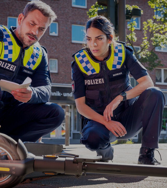 "Notruf Hafenkante - Neues Leben": Der Polizist Kris Freiberg (Marc Barthel) und seine Kollegin Isa Nowak (Lilli Hollunder) sitzen nebeneinander in der Hocke auf einem Gehweg. Vor ihnen liegt ein Elektroroller, den sie sich beide ansehen.