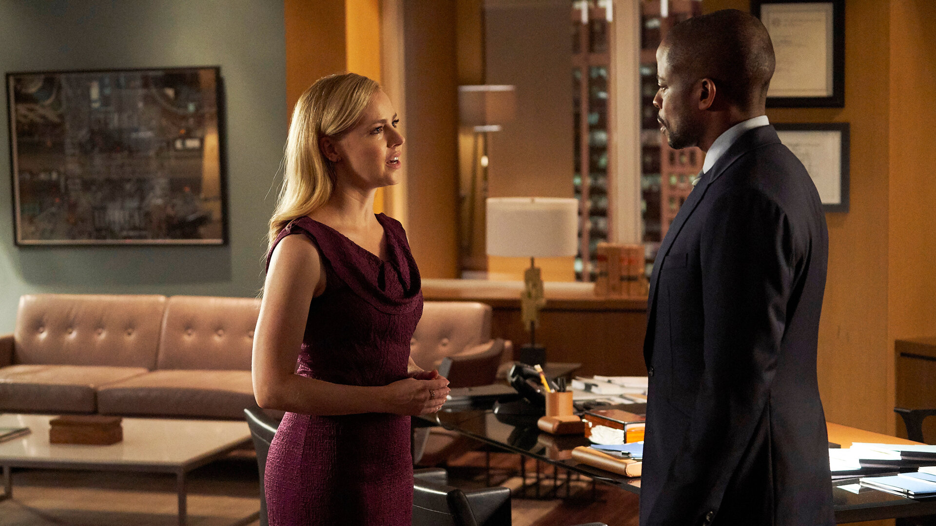"Suits - Rocky 8": Katrina (Amanda Schull), die ein bordeauxfarbenes Kleid trägt, steht Alex (Dulé Hill)  - im dunklen Anzug - gegenüber. Im Hintergrund ist das elegante, in warmen Brauntönen gehaltene Büromobiliar zu erkennen sowie einige gerahmte Diplome an der Wand.