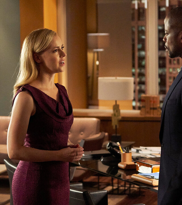 "Suits - Rocky 8": Katrina (Amanda Schull), die ein bordeauxfarbenes Kleid trägt, steht Alex (Dulé Hill)  - im dunklen Anzug - gegenüber. Im Hintergrund ist das elegante, in warmen Brauntönen gehaltene Büromobiliar zu erkennen sowie einige gerahmte Diplome an der Wand.