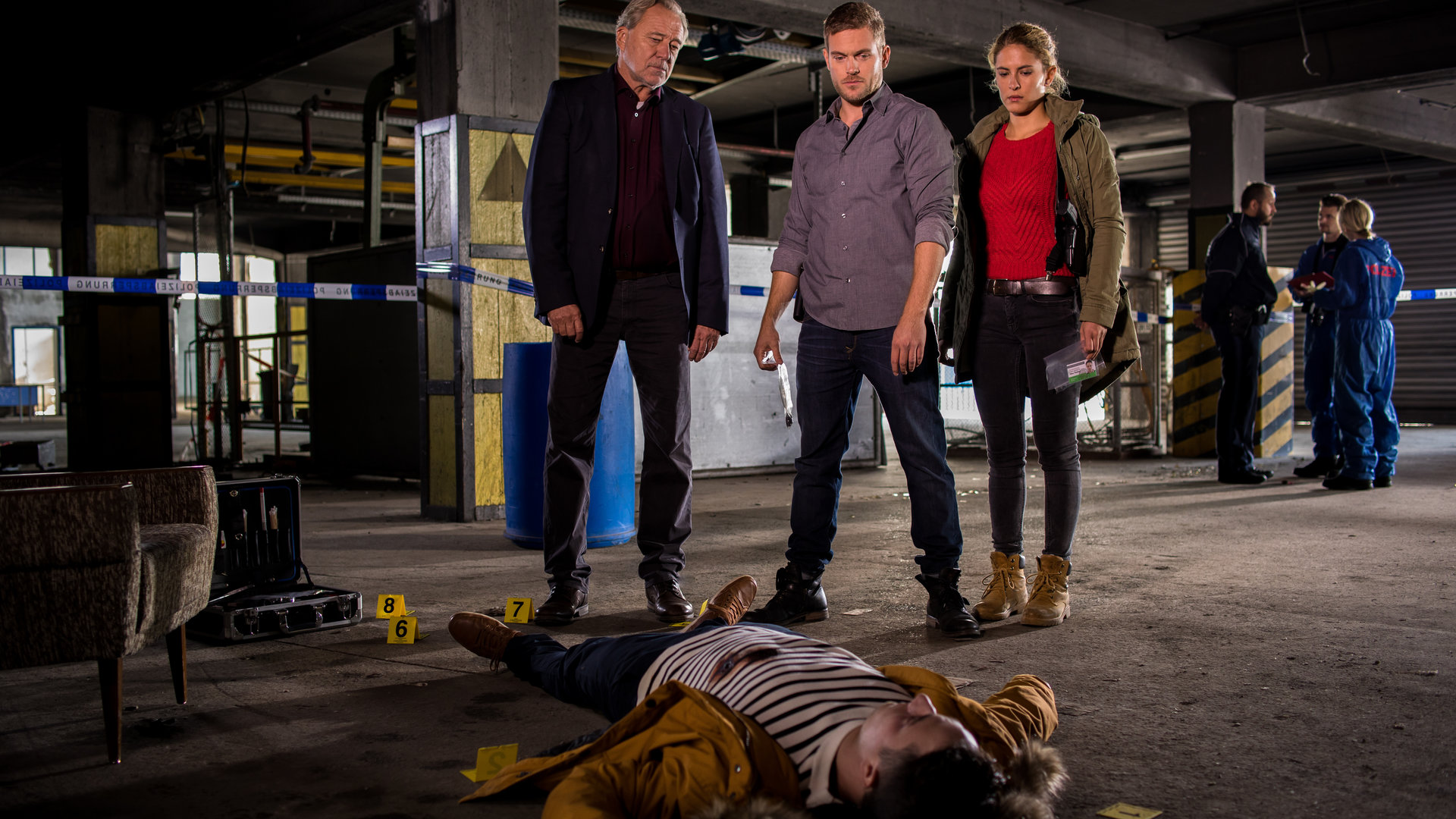 "SOKO München - Die Verlorenen": In einem Fabrikgebäude liegt eine männliche Leiche auf dem Boden. Arthur Bauer (Gerd Silberbauer), Dominik Morgenstern (Joscha Kiefer) und Toni Bischoff (Amanda da Gloria) stehen hinter der Leiche und schauen auf sie. Im Hintergrund sind Kollegen von der Spusi (Komparsen) zu sehen.