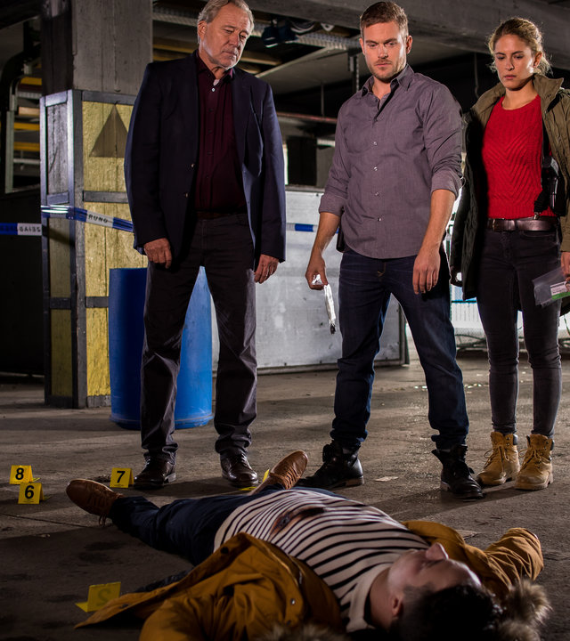 "SOKO München - Die Verlorenen": In einem Fabrikgebäude liegt eine männliche Leiche auf dem Boden. Arthur Bauer (Gerd Silberbauer), Dominik Morgenstern (Joscha Kiefer) und Toni Bischoff (Amanda da Gloria) stehen hinter der Leiche und schauen auf sie. Im Hintergrund sind Kollegen von der Spusi (Komparsen) zu sehen.