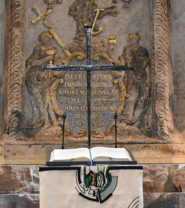 "Festlicher Neujahrsgottesdienst - Probieren wir's mit Liebe": Über einer aufgeschlagenen Bibel steht das Nagelkreuz in der Dresdner Frauenkirche. Es ist aus dunkelgrauem Metall geschmiedet. Links und rechts davon stehen jeweils zwei hohe Kerzenständer, ebenfalls aus dunklem Metall, mit weißen Kerzen.
