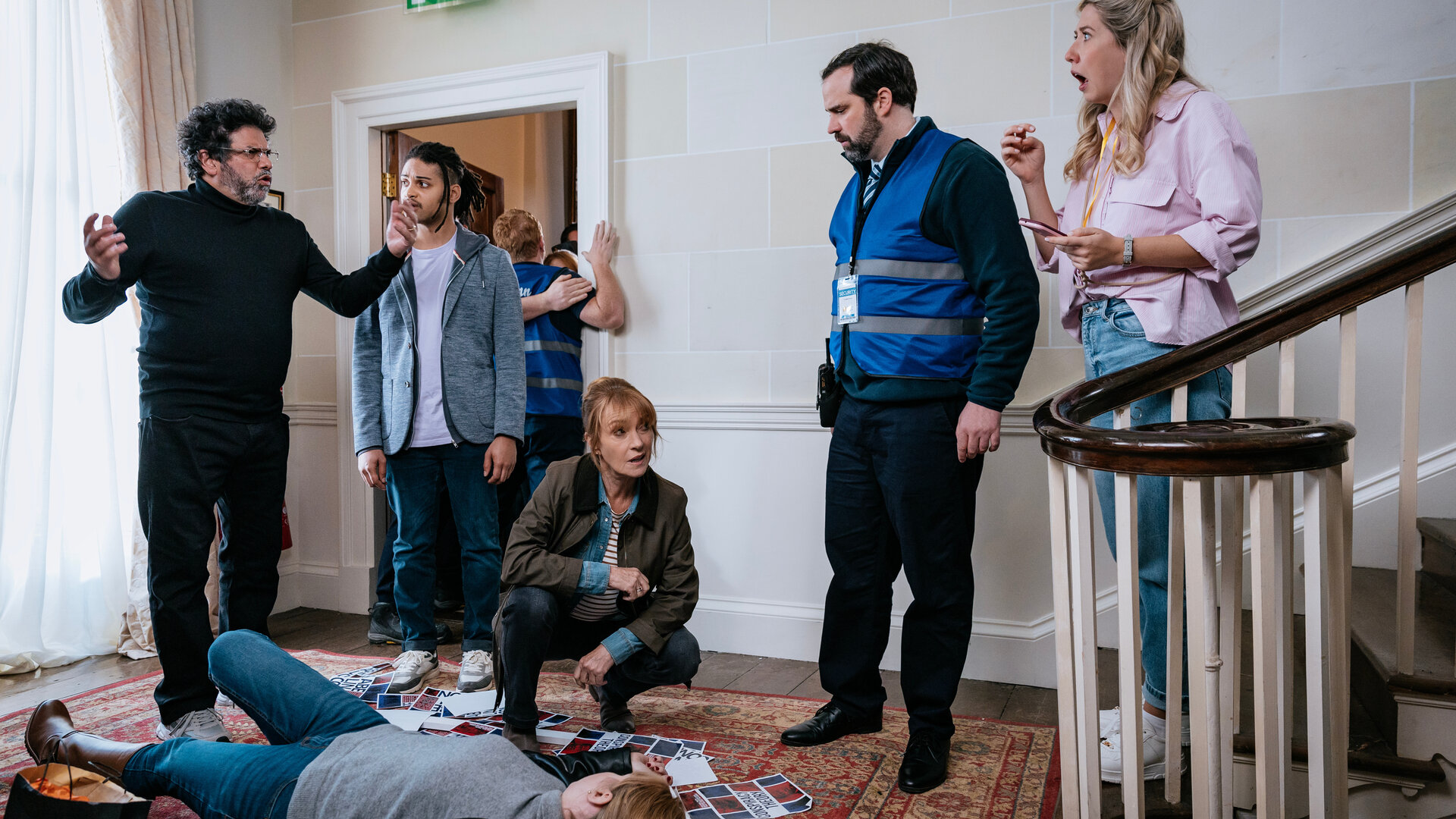 "Harry Wild – Mörderjagd in Dublin - Jemand hat mich umgebracht!": Harry Wild (Jane Seymour) nimmt kniend die tote Tina Mulcahy (Aileen Mythen) in Augenschein. Die Leiche liegt vor einer Treppe auf einem Teppich. Um sie herum sind Werbeflyer verstreut. Glenn Talbot (Paul Tylak) steht dabei und gestikuliert. Die Convention-Veranstalterin Mary O'Driscoll (Ali Hardiman) wirkt erschrocken. Fergus Reid (Rohan Nedd) und Security-Chef Ollie Reilly (Colm O'Brien) beobachten die Szene.