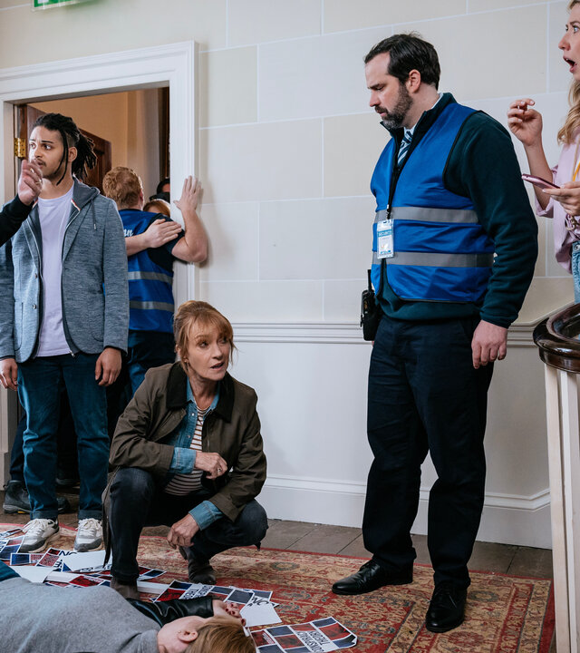 "Harry Wild – Mörderjagd in Dublin - Jemand hat mich umgebracht!": Harry Wild (Jane Seymour) nimmt kniend die tote Tina Mulcahy (Aileen Mythen) in Augenschein. Die Leiche liegt vor einer Treppe auf einem Teppich. Um sie herum sind Werbeflyer verstreut. Glenn Talbot (Paul Tylak) steht dabei und gestikuliert. Die Convention-Veranstalterin Mary O'Driscoll (Ali Hardiman) wirkt erschrocken. Fergus Reid (Rohan Nedd) und Security-Chef Ollie Reilly (Colm O'Brien) beobachten die Szene.