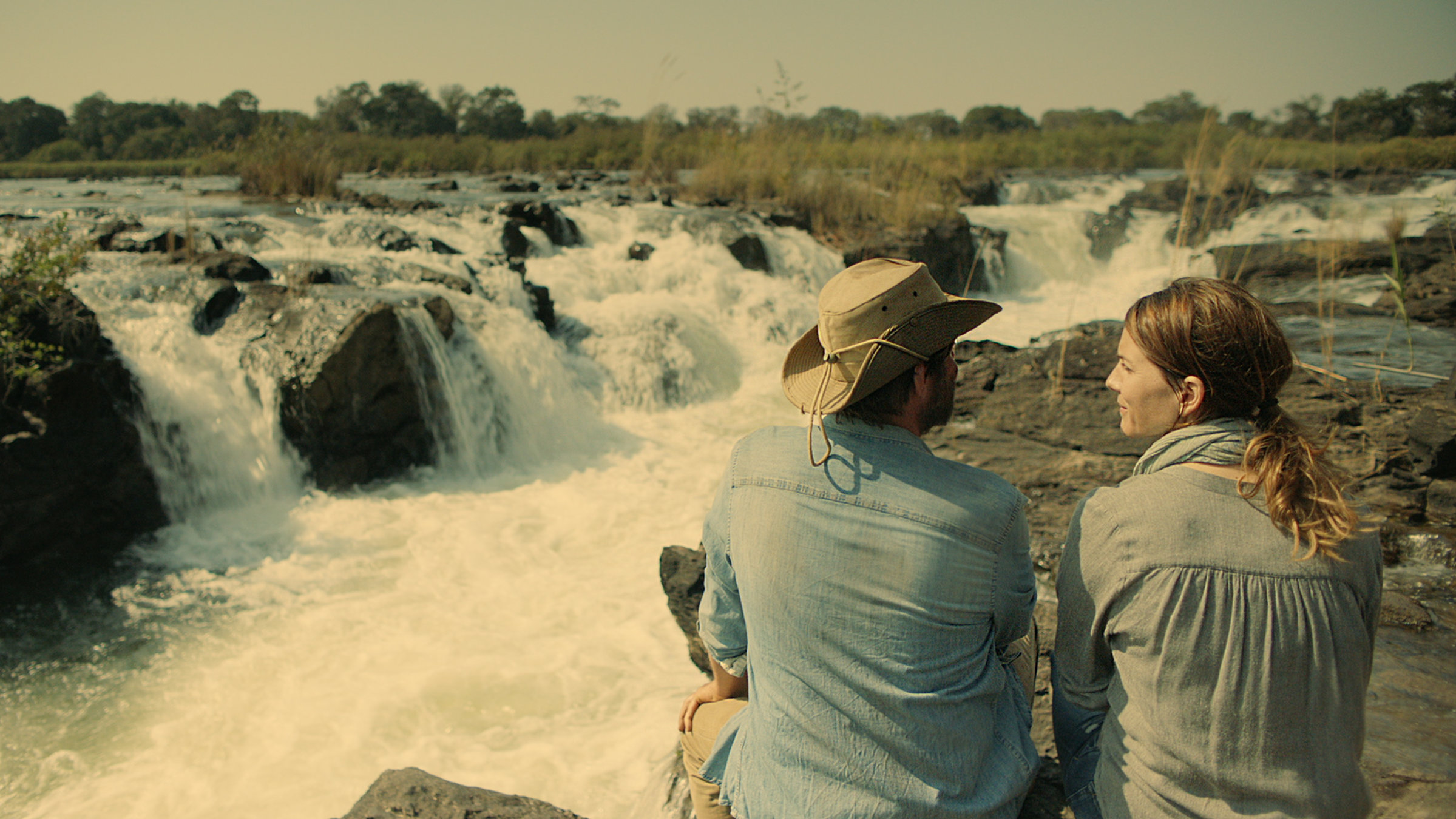 "Fluss des Lebens - Okavango, Fremder Vater": Gunnar (Roeland Wiesnekker) und Charlotte (Christina Hecke) sitzen vor einem Wasserfall des Okavango. Die beiden sind von hinten zu sehen. Charlotte schaut Gunnar an, der nach vorne schaut.