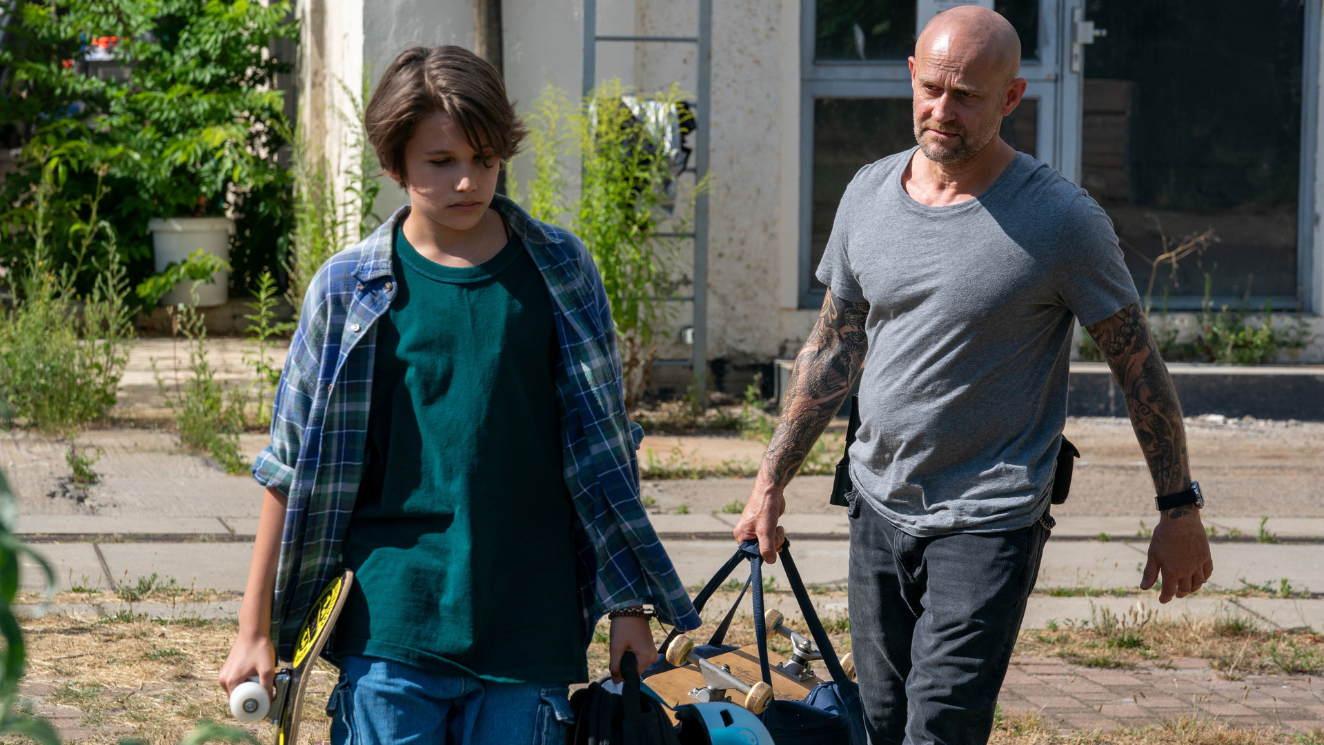 "Jenseits der Spree - Tommy": Tommy (Linus Kranich) und Robert (Jürgen Vogel) tragen Skateboards und Taschen, während sie über ein Gelände laufen. Robert geht knapp hinter Tommy und schaut ihn dabei fragend an.