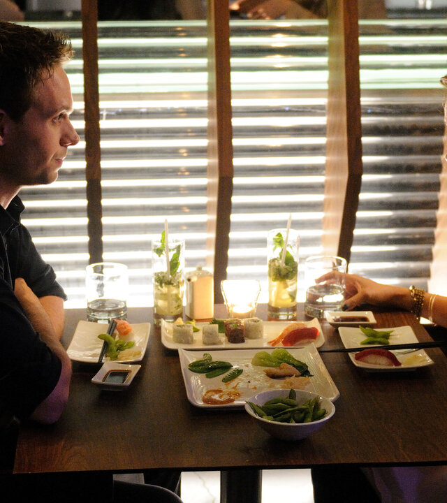 "Suits - Alleingänge": Mike (Patrick J. Adams) und Rachel (Meghan Markle) sitzen sich bei gedimmten Licht in einem modernen Restaurant gegenüber. Auf dem Tisch stehen Sushi, Edamame und Cocktails, beide wirken nachdenklich und aufmerksam.