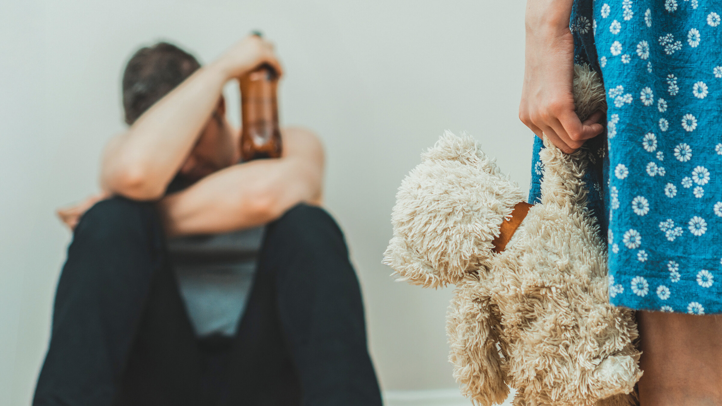 "37°: Vergiftete Kindheit - Wenn Alkohol Familien belastet": Ein Mann sitzt am Boden. Er hat die Arme auf den Knien abgelegt, den Kopf nach unten gesengt. In der einen Hand hält er eine Bierflasche. Neben ihm steht ein Kind in einem blauweiß geblümten Kleid. Sie hält einen Teddy in der Hand.