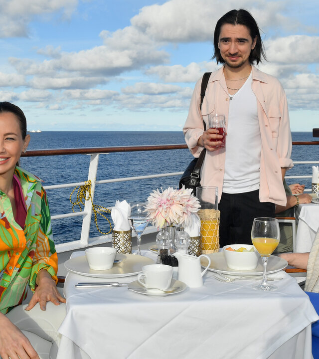 "Das Traumschiff: Argentinien": Auf dem Schiffsdeck: Glücklich lächelnd sitzen Cora Bruns (Friederike Linke) und die Yogalehrerin (Alessandra Meyer-Wölden) am gedeckten Frühstückstisch. Tobias Bender (Ivo Kortlang) steht am Tisch und hält ein gefülltes Glas in den Händen.