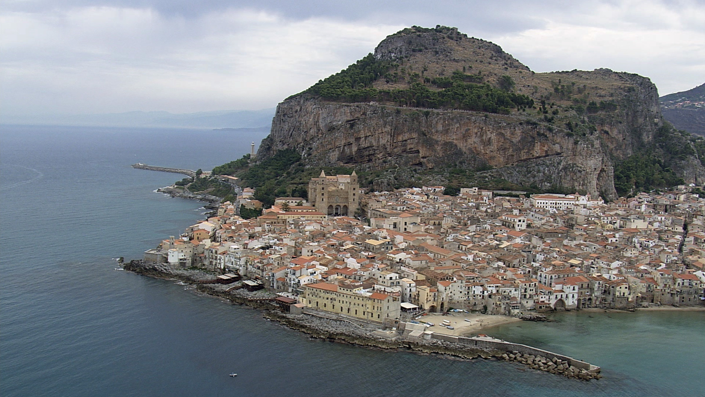 "Süditalien von oben": Die Küstenstadt Cefalù auf Sizilien mit großem Kirchengebäude vor einem Berg mit einer steilen Felswand.
