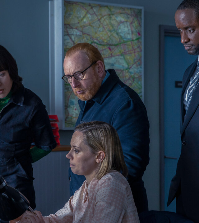 "The Chelsea Detective - Mord aus Leidenschaft": DC Jess Lombard (Lucy Phelps) sitzt am Schreibtisch vor einem PC. DS Layla Walsh (Vanessa Emme), DI Max Arnold (Adrian Scarborough) und DC Connor Pollock (Peter Bankolé) stehen hinter ihr und blicken ebenfalls auf den Bildschirm.