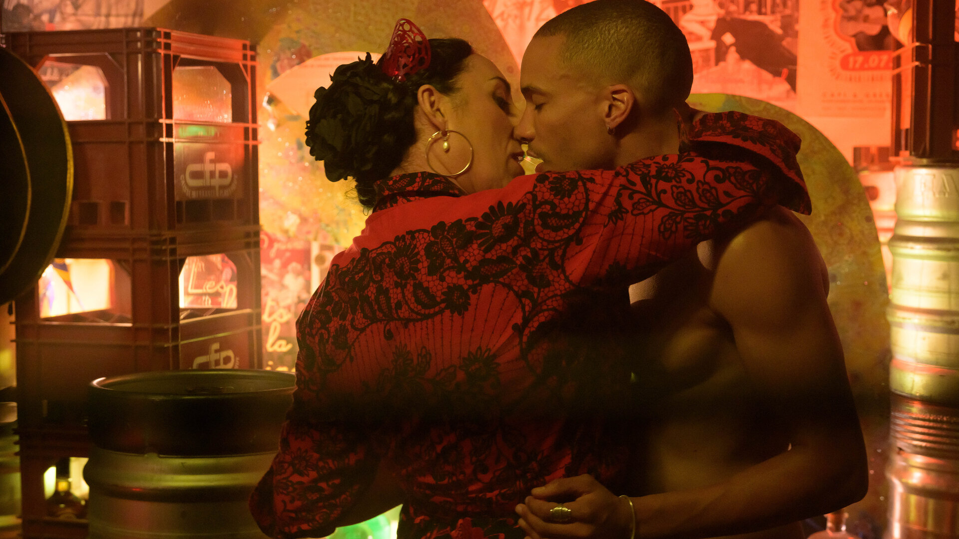 "Escort Boys - Besondere Bedürfnisse": Mimi (Rossy de Palma) in rot gekleidet, die schwarzen Haare zu einem Dutt gebunden, legt ihren Arm um Zak (Corentin Fila), dessen Oberkörper nackt ist. Ihre Gesichter nähern sich dabei an, für einen Kuss.