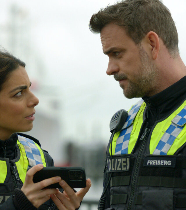 "Notruf Hafenkante - Im Schock": Nahaufnahme: Polizistin Isa Nowak (Lilli Hollunder) und ihr Kollegen Kris Freiberg (Marc Barthel) stehen nebeneinander im Freien und sehen sich an. Isa zeigt Kris etwas auf ihrem Handy.