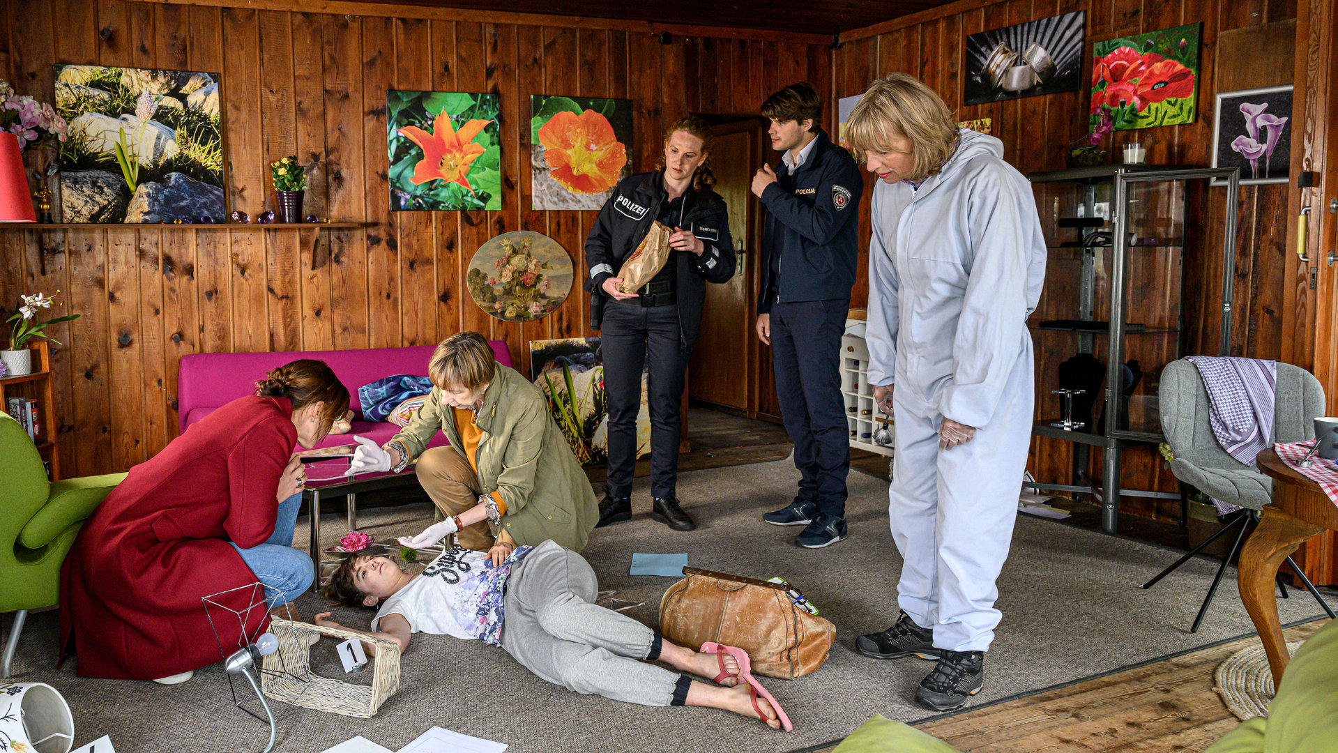 "SOKO Wismar – Brötchen holen": In ihrem Wohnzimmer liegt die junge Goldschmiedin Kim Olemacher (Tamara Spieth). Dr. Helene Sturbeck (Katharina Blaschke) und Kommissarin Karoline Joost (Nike Fuhrmann) knien über ihr. Paula Moorkamp (Stella Hinrichs), Eddi Jansons (Gustavs Gailus) und Roswitha Prinzler (Silke Matthias) schauen herab.