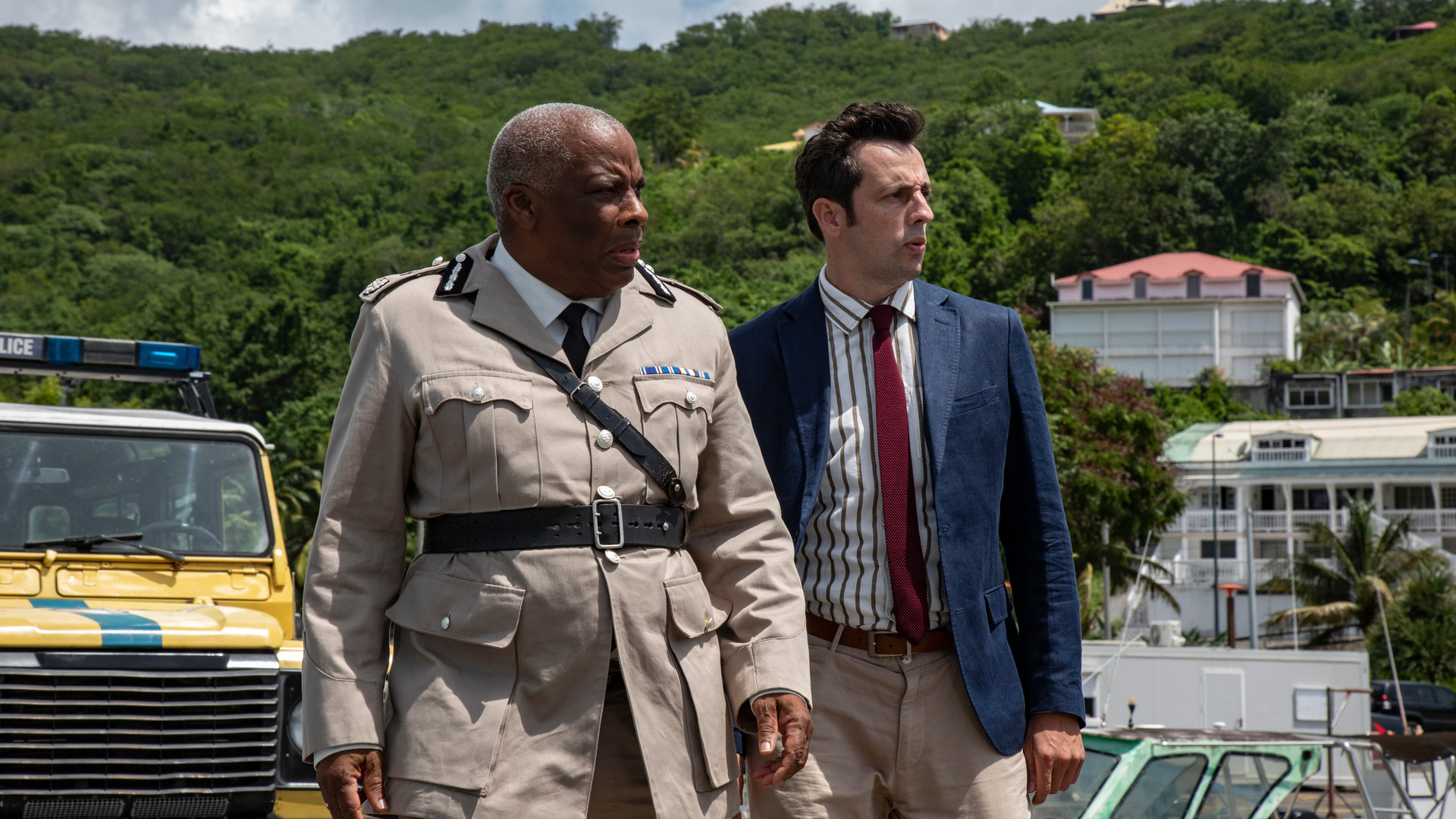 "Death in Paradise - Weihnachten unter Palmen 2": DI Parker (Ralf Little) und Commissioner Patterson (Don Warrington) stehen nebeneinander vor einem Streifenwagen und schauen konzentriert in die Ferne.