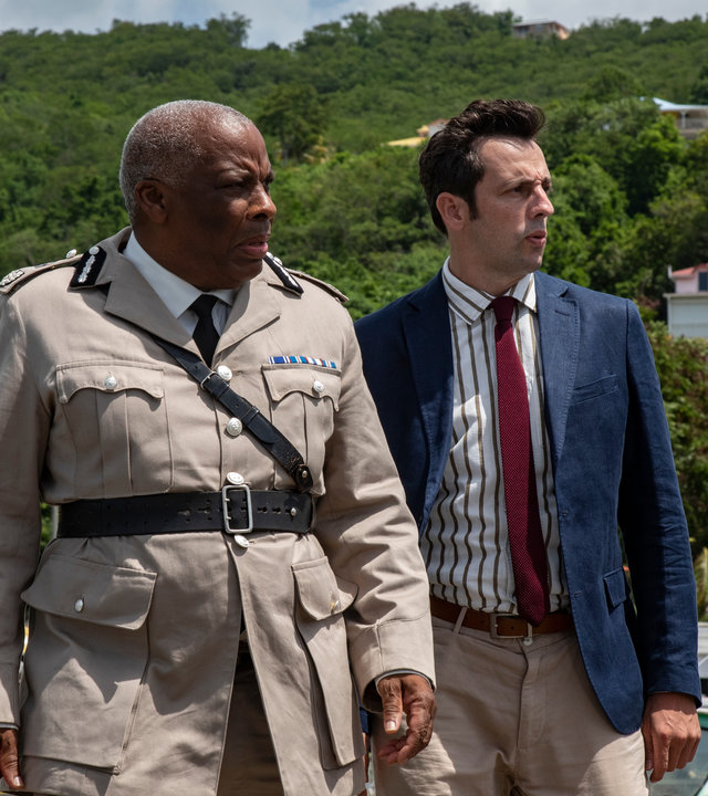 "Death in Paradise - Weihnachten unter Palmen 2": DI Parker (Ralf Little) und Commissioner Patterson (Don Warrington) stehen nebeneinander vor einem Streifenwagen und schauen konzentriert in die Ferne.