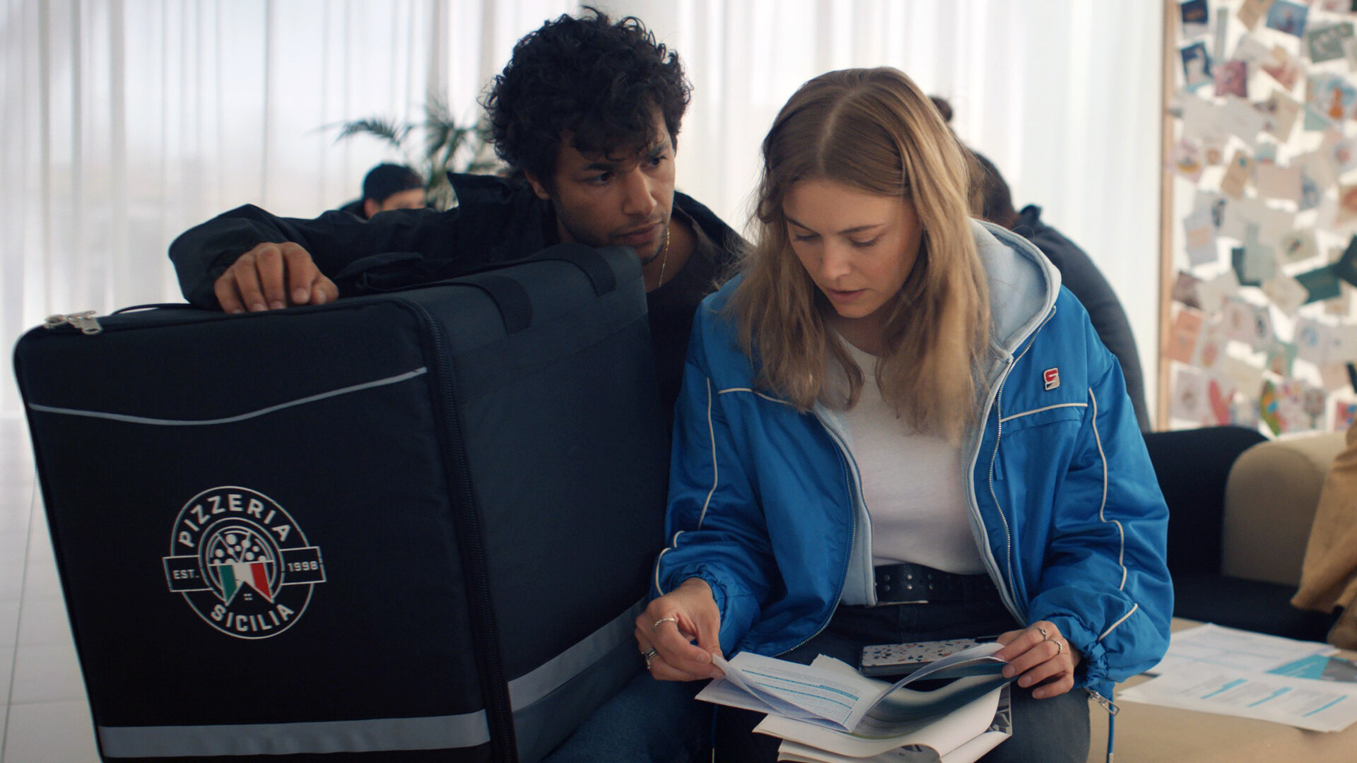 "The Club - Der Jetzt-oder-nie-Club": Im Wartezimmer sitzen Charlie (Roos Dickmann) und Ziggy (Achraf Koutet) nebeneinander. Charlie hält eine Broschüre in den Händen, während Ziggy die Pizza-Liefertasche auf seinem Schoß hält und Charlie dabei ansieht.