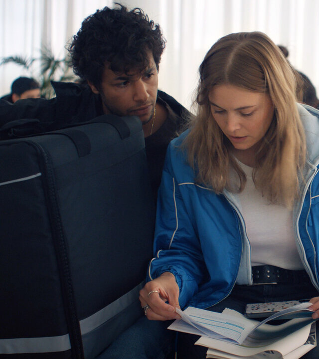 "The Club - Der Jetzt-oder-nie-Club": Im Wartezimmer sitzen Charlie (Roos Dickmann) und Ziggy (Achraf Koutet) nebeneinander. Charlie hält eine Broschüre in den Händen, während Ziggy die Pizza-Liefertasche auf seinem Schoß hält und Charlie dabei ansieht.