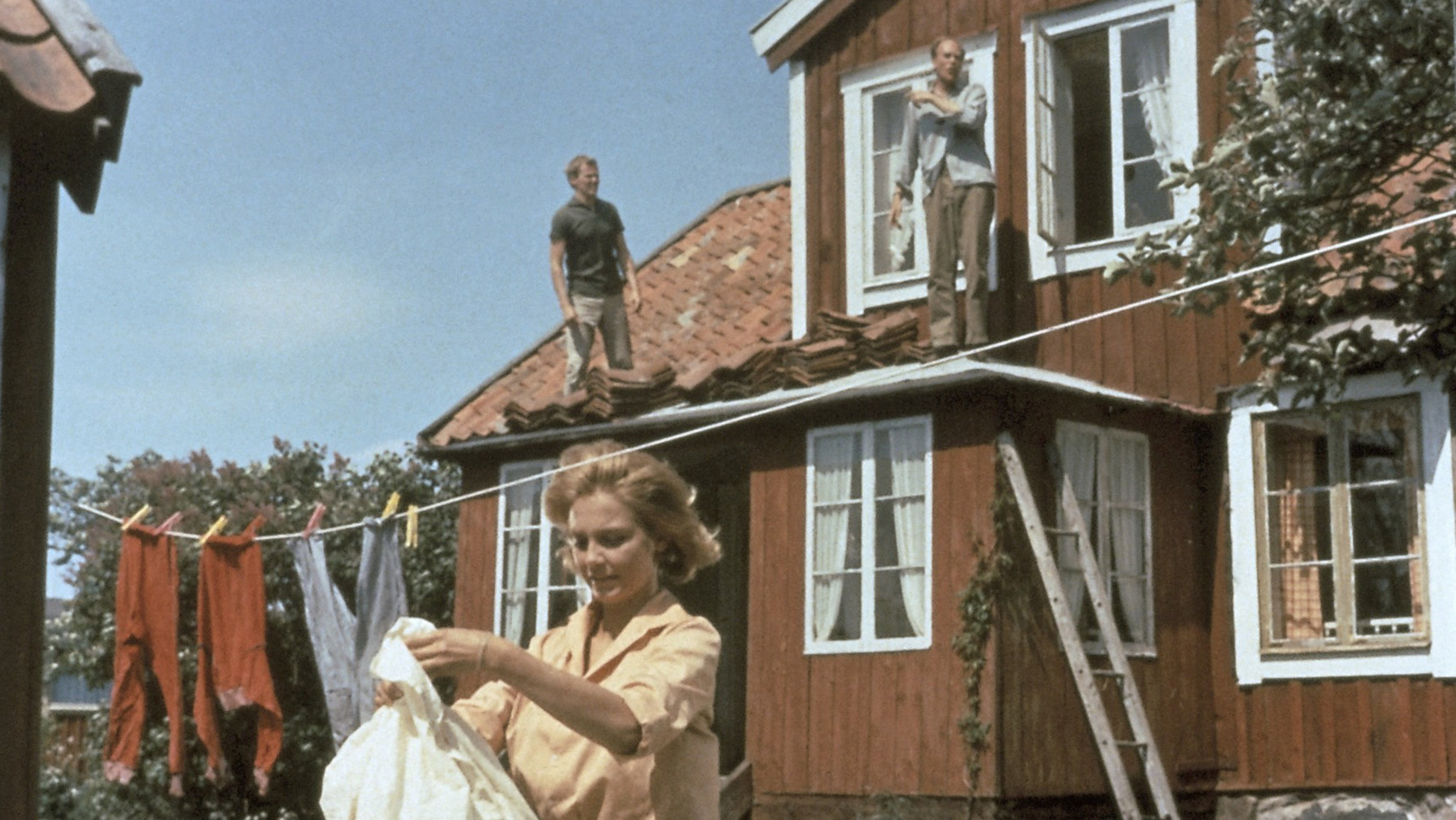 "Ferien auf Saltkrokan - Das Trollkind": Eine Frau hängt vor einem Holzhaus Wäsche auf. Im Hintergrund sieht man zwei Männer auf einem Vordach stehen.