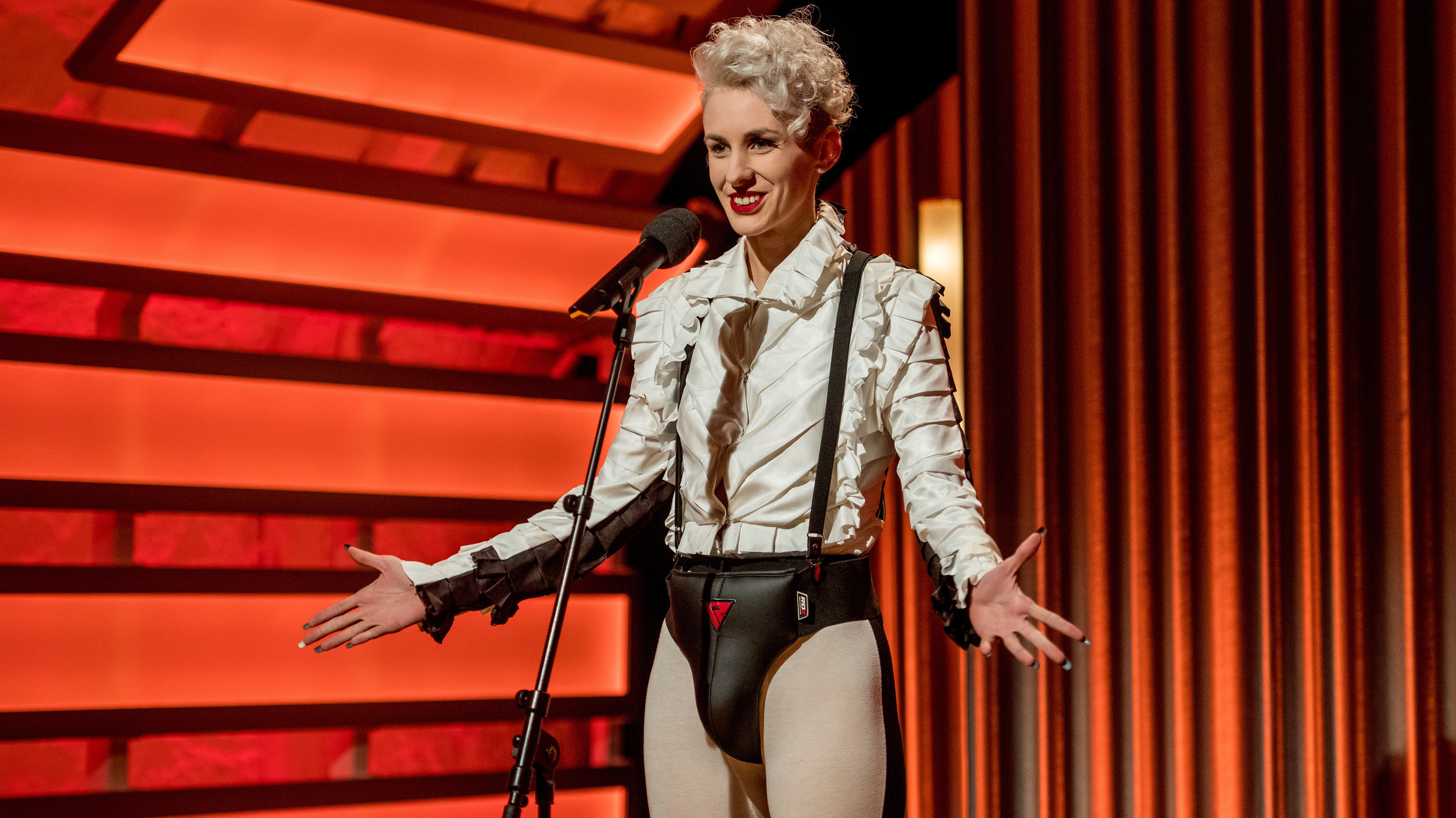 "Lisa Eckhart: Als ob Sie Besseres zu tun hätten": Lisa Eckhart steht vor einem Mikrofon auf der Bühne. Sie trägt eine schwarz-weiße Rüschchenbluse, graue Leggins und eine schwarze kurze vorne gepolsterte Hose. Sie hat blond gelocktes Haar. Eine Seite von ihrem Kopf ist rasiert.