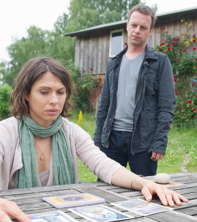 "SOKO Wismar - Schlechte Karten für Belinda": Silke Jaspers (Jana Pallaske) sitzt in einem Garten an einem Holztisch und liest in den Karten. Sven Herzog (Michael Härle) steht hinter ihr und beobachtet sie.