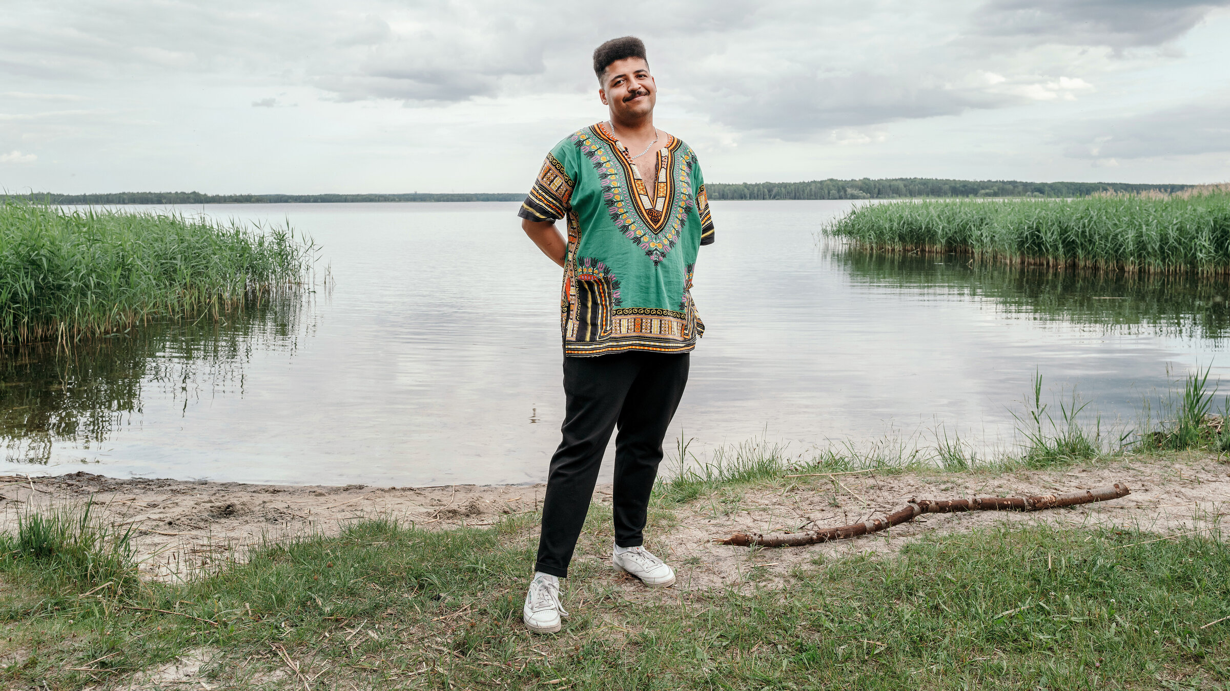 "37°Leben: Mein Erbe der Einheit": Dennis, ein junger Mann mit dunklem Teint, steht am Ufer eines ruhigen Sees, der Himmel ist größtenteils bewölkt. Er steht auf einer kleinen Grasfläche, die Hände sind locker in die Hosentaschen gesteckt, die Körperhaltung wirkt entspannt und freundlich.