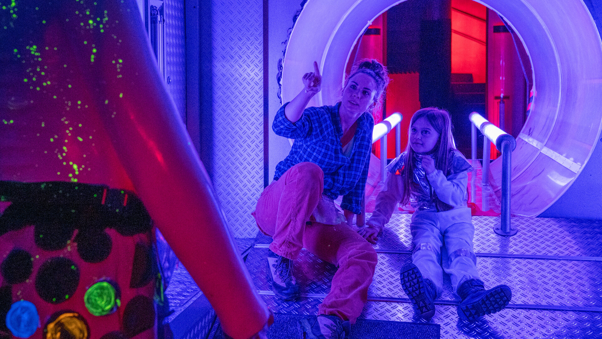 "SOKO Stuttgart - Tod im Wunderland": Emily Hellwig (Julia E. Lenska) und ihre Tochter Mia (Komparsin) sitzen in einem Indoor-Spielplatz in einem Raum, der einem Raumschiff nachempfunden ist. Das Licht ist bläulich. Emily zeigt auf eine glitzernde und leuchtende Figur, die vor ihnen steht, und Mia schaut interessiert hin.