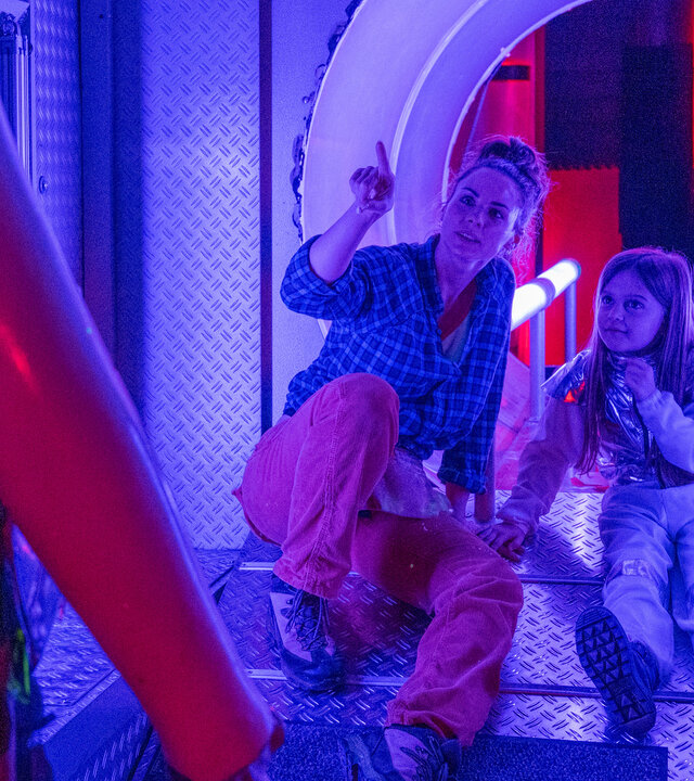 "SOKO Stuttgart - Tod im Wunderland": Emily Hellwig (Julia E. Lenska) und ihre Tochter Mia (Komparsin) sitzen in einem Indoor-Spielplatz in einem Raum, der einem Raumschiff nachempfunden ist. Das Licht ist bläulich. Emily zeigt auf eine glitzernde und leuchtende Figur, die vor ihnen steht, und Mia schaut interessiert hin.
