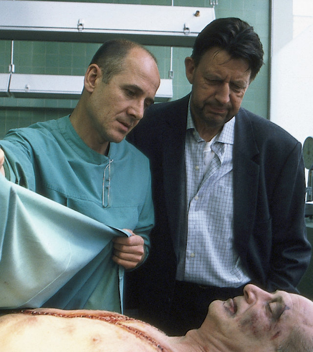 "Der letzte Zeuge - Ich hasse meine Mutter": Dr. Robert Kolmaar (Ulrich Mühe) und Joe Hoffer (Jörg Gudzuhn) stehen vor der Leiche von David Taylor (Matthew Burton) am Obduktionstisch und begutachten sie.