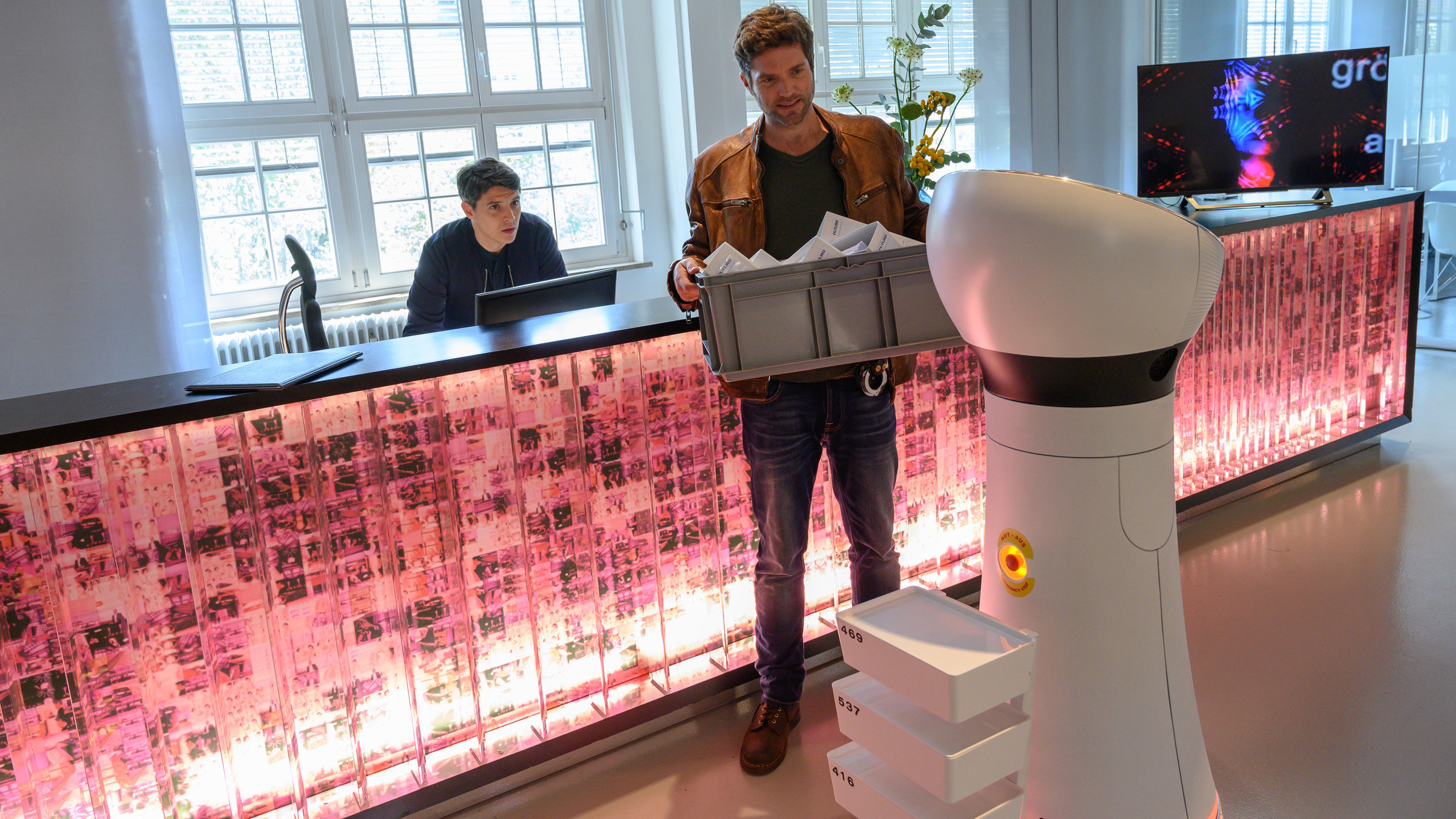 “SOKO Stuttgart – Hotel Futuro“: Im Foyer des modernen Boutique-Hotels „Hotel Futuro“ steht Rico Sander (Benjamin Strecker) am Computer hinter dem Empfangstresen der Rezeption. Auf dem Tresen steht ein Bildschirm auf dem eine Videoinstallation läuft. Jo Stoll (Peter Ketnath) steht vor dem Tresen, und hält eine graue Kiste mit Ordnern in den Händen. Vor ihm steht ein weißer Service-Roboter, den beide Ermittler kritisch beäugen.