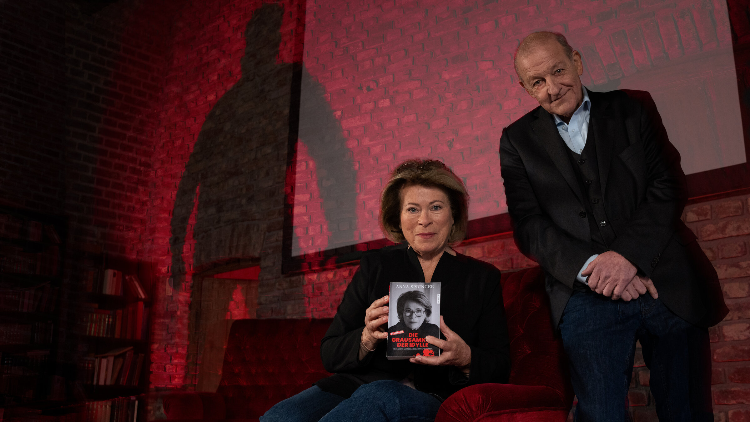 "Wilsberg - Blut geleckt": Anna Springer (Rita Russek) sitzt in einem großen roten Samtsessel und präsentiert vorsichtig lächelnd ihr "True Crime"-Buch "Die Grausamkeit der Idylle", welches sie in den Händen hält. Wilsberg (Leonard Lansink) sitzt auf der Lehne des Sessels und blickt ebenfalls lächelnd in die Kamera. Im Hintergrund erhebt sich der Schatten einer unbekannten Person an der Wand, der für Anna Springer und Wilsberg gefährlich wirkt.