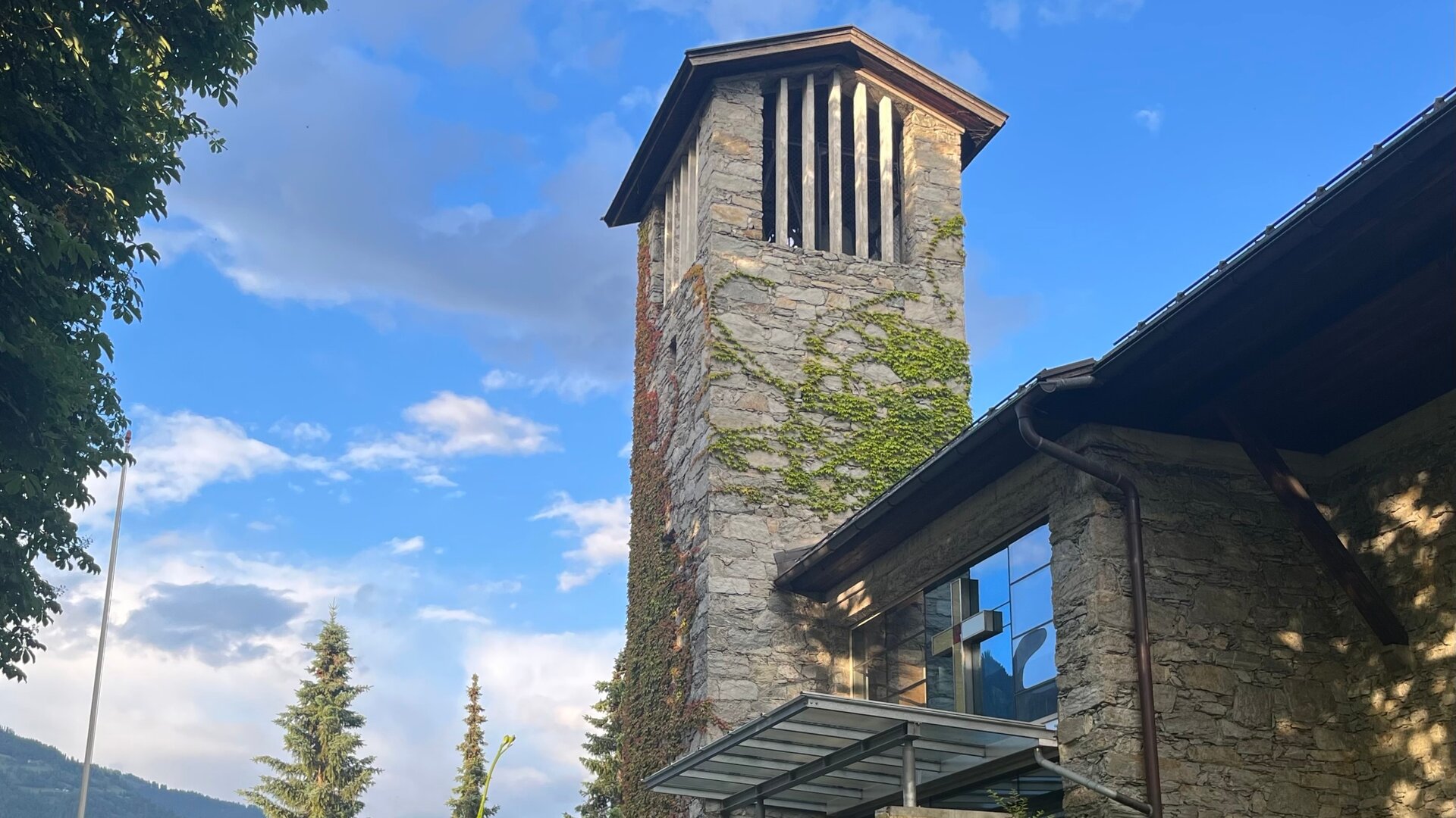 "Evangelischer Gottesdienst - Dem Himmel so nah": Der Turm, der aus grauem Stein gebauten Kirche auf der Emberger Alm, ragt in den blauen Himmel mit weißen Wolken. Neben dem Turm befindet sich ein Ausschnitt des Kirchengebäudes, auf der anderen Seite stehen Bäume vor Hügeln.