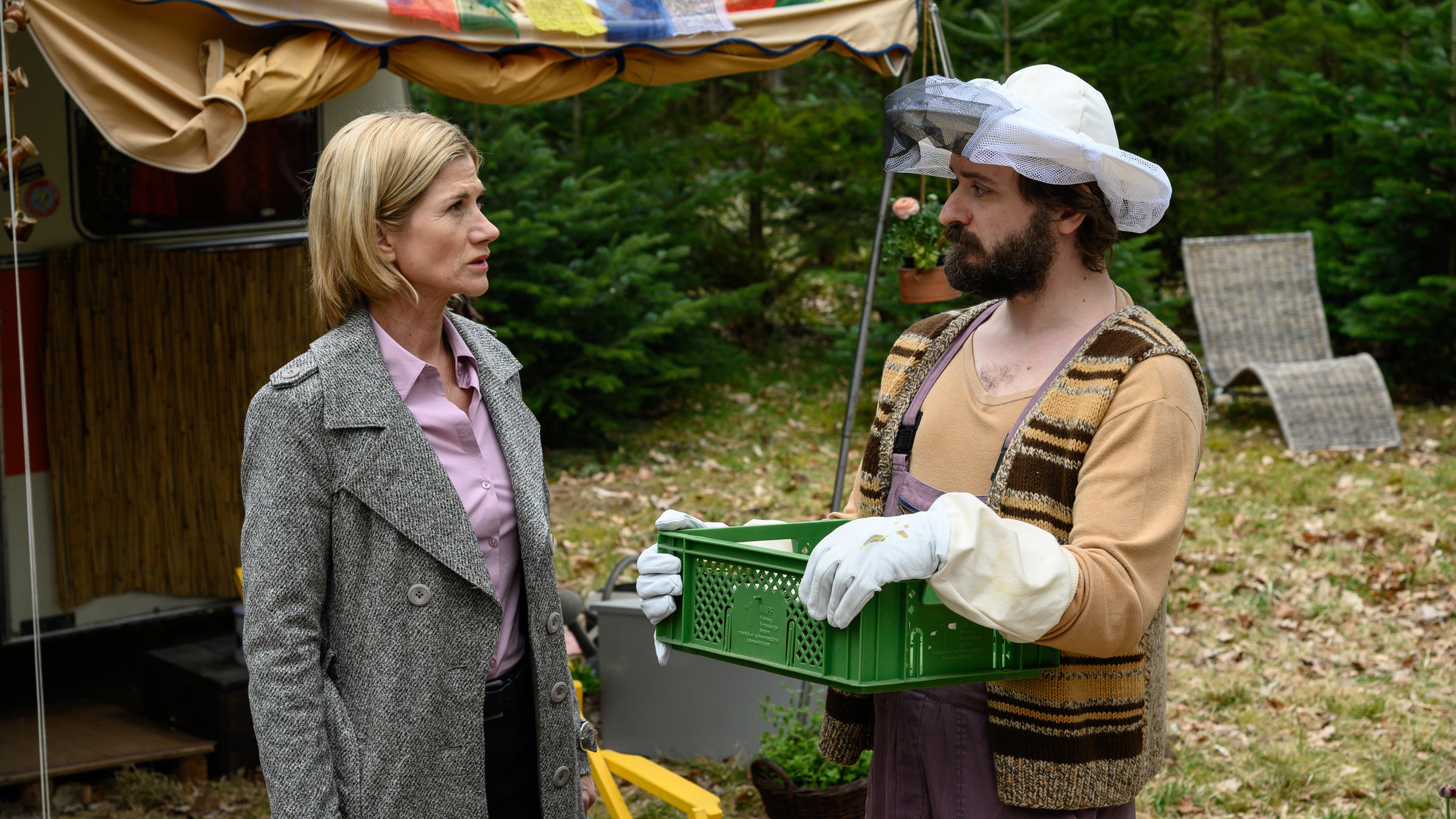 "SOKO Stuttgart - Der Bienenstock": Martina Seiffert (Astrid M. Fünderich) steht vor einem Bauwagen mit Vorzelt und spricht mit Bernhard „Berni“ Lindau (Daniel Christensen), der einen Imkerhut und –handschuhe trägt.